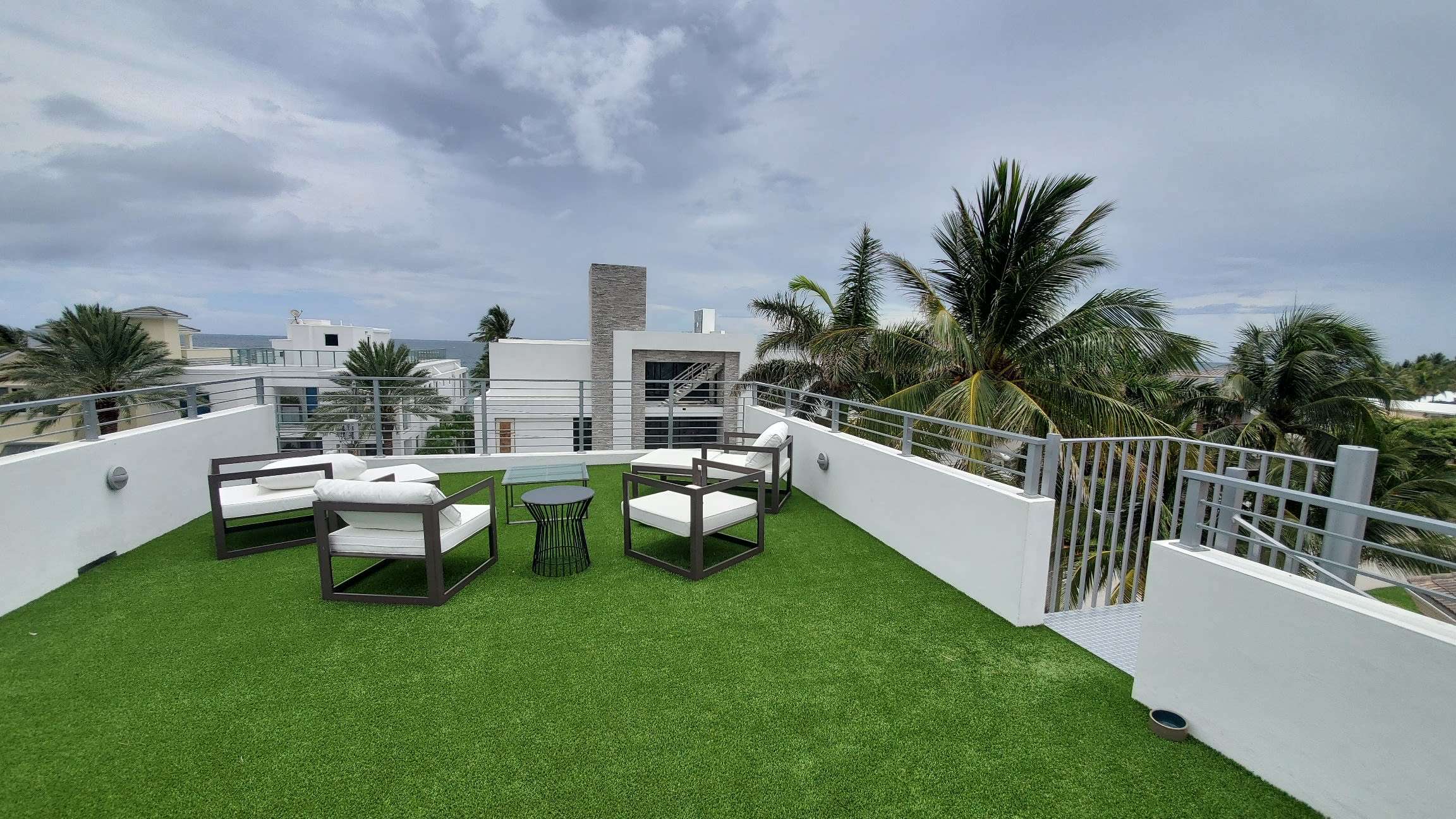Rooftop patio with artificial grass flooring, featuring modern outdoor furniture with a sofa and chairs, bordered by a white railing with tropical palm trees and a view of overcast skies in the background.