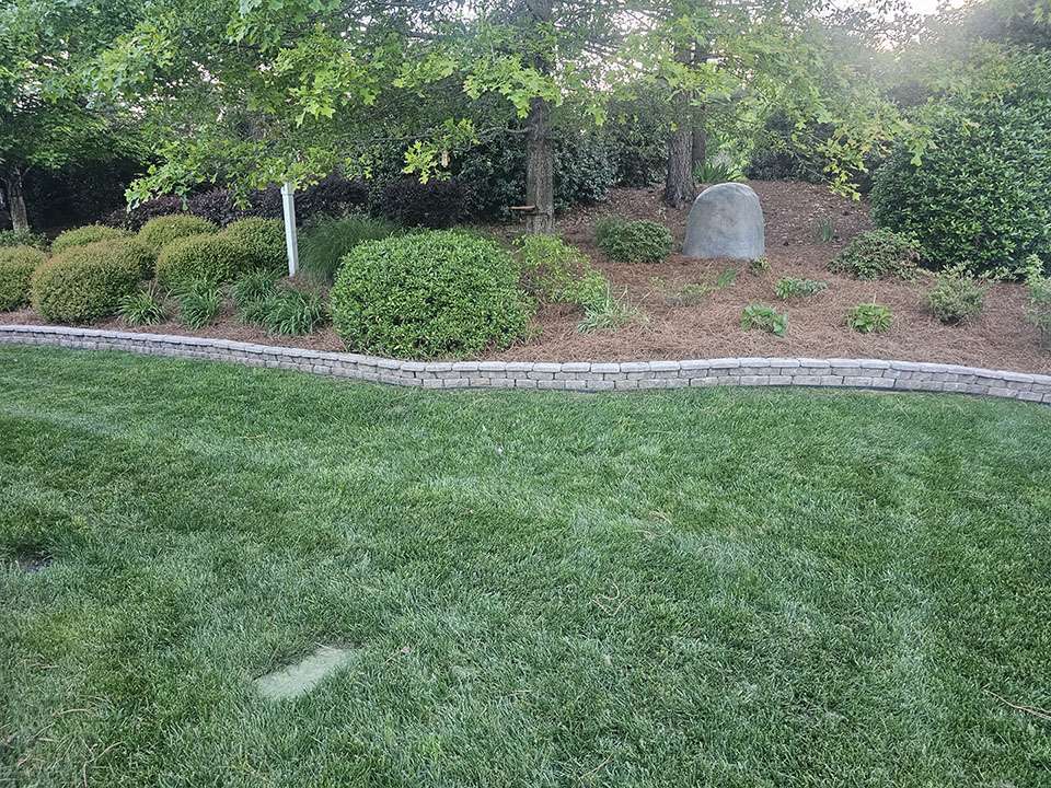 Lush lawn leading to a hardscape retaining wall made of stacked stones, bordering a garden with various shrubs and a central boulder, under the shade of mature trees.