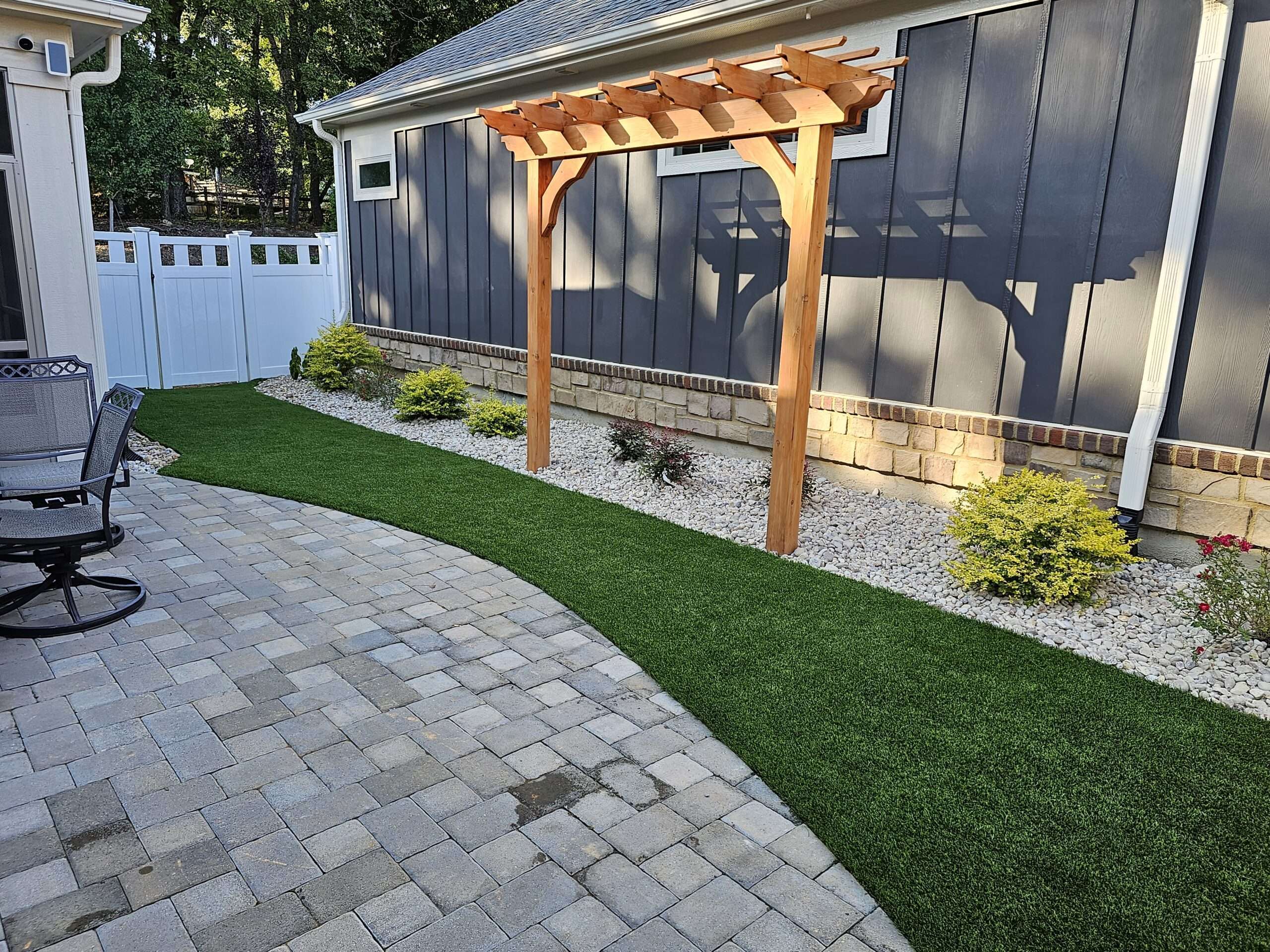 Neatly designed backyard with a paver patio leading to a pergola, bordered by vibrant artificial turf and landscaping rocks with flowering shrubs alongside a grey house with white trim.